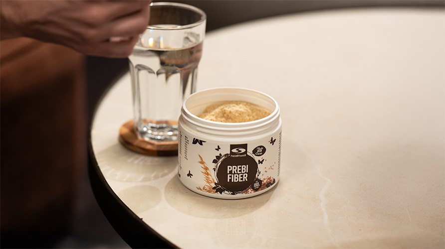 Healthwell PrebiFiber og et glas vand p et bord, en hnd er klar til at blande.